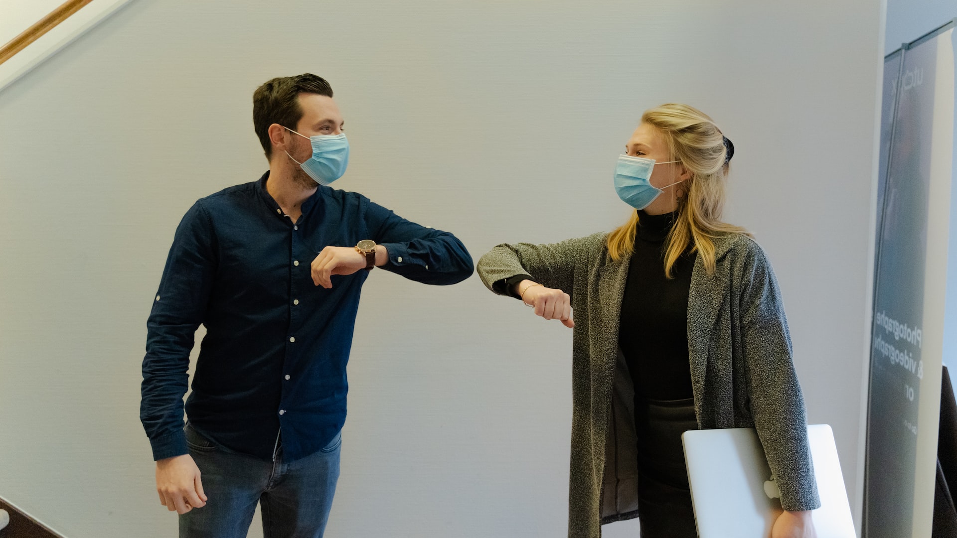A woman and a man, both wearing a surgical mask, perform an elbow greeting. Photo by Maxime on Unsplash.