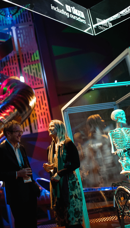 A man in a suit stands holding a drink next to a woman in a black jacket and scarf, in the background is a museum exhibit. The scene is illuminated by colourful lighting. Behind them, a display features a human skeleton.