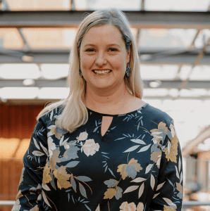 Professor Kathryn MacCallum wearing a floral black dress stands in a well-lit area with a visible wooden background