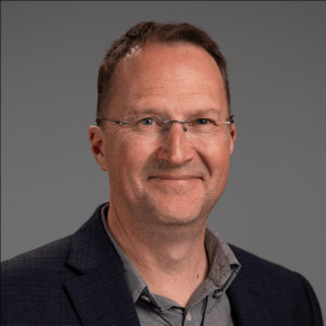 Dr. Mark Nichols, a middle-aged man with short brown hair and glasses is smiling while wearing a dark blazer over a light shirt