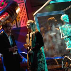 Two women and a man stand in a dark exhibition space beside a glowing skeleton display, with neon-lit panels and an illuminated ceiling installation overhead.