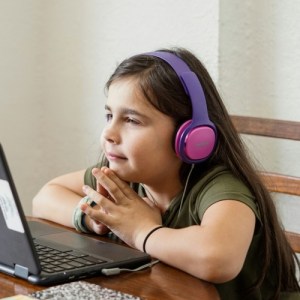 Young cute primary-school aged girl with headphones on looking at a laptop while participating and learning in a remote virtual learning class